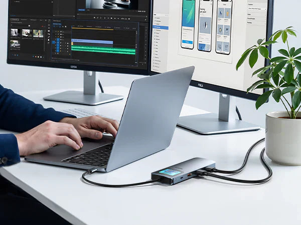 Person typing on a laptop connected to a multi-port USB-C hub, which extends the display to a large desktop monitor showing design software.